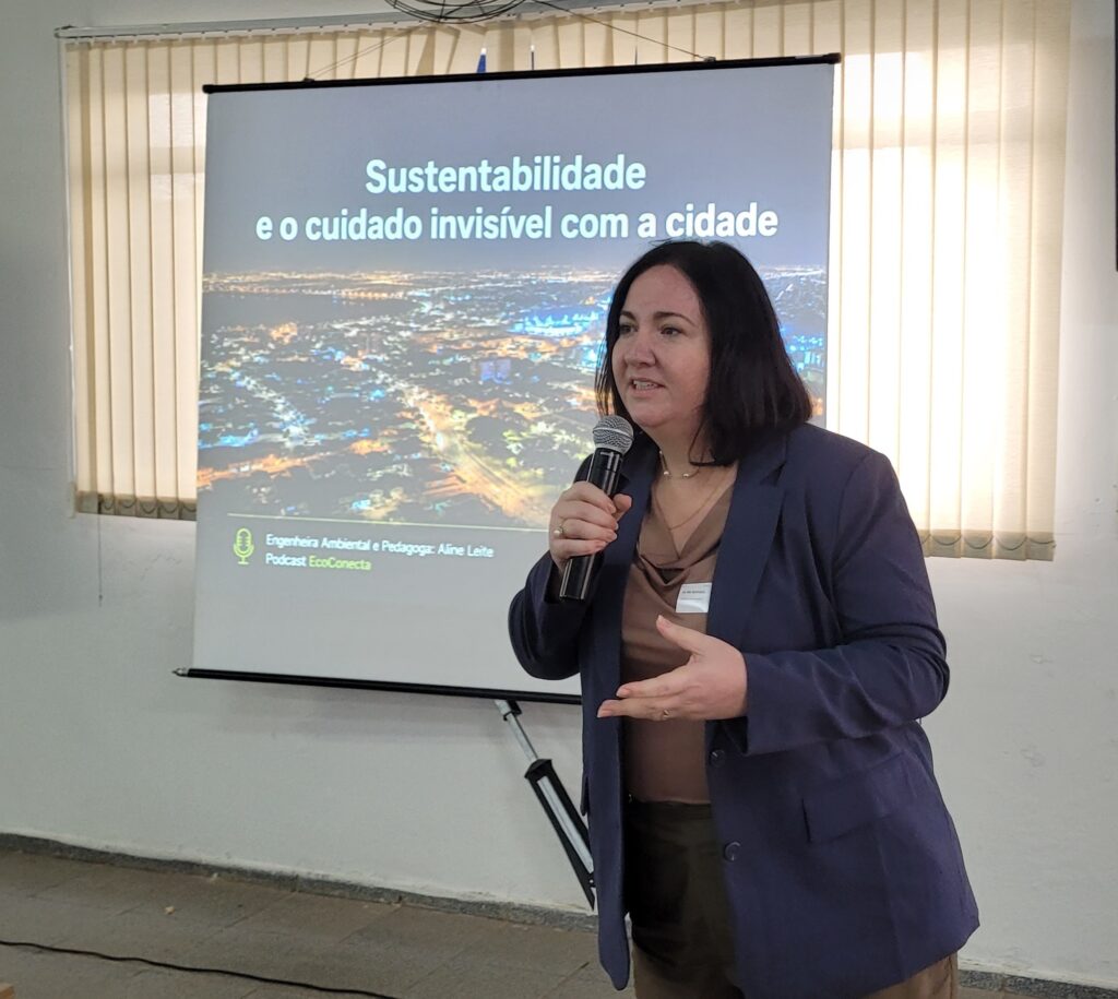 A imagem mostra uma mulher apresentando em um ambiente interno, possivelmente uma sala de conferências ou reunião. Ela está em pé, segurando um microfone com a mão direita e gesticulando com a mão esquerda. A mulher tem cabelos escuros e está vestindo um blazer azul escuro sobre uma blusa marrom. Ela também usa um crachá de identificação preso ao blazer. Ao fundo, há uma tela de projeção que exibe o título da apresentação: "Sustentabilidade e o cuidado invisível com a cidade". A apresentação é feita pela "Engenheira Ambiental e Pedagoga: Aline Leite" e está associada ao "Podcast EcoConecta". A imagem na tela mostra uma vista aérea noturna de uma cidade iluminada, destacando as ruas e avenidas com luzes amarelas. O ambiente é claro, com paredes brancas e janelas cobertas por persianas verticais. A iluminação é artificial, garantindo boa visibilidade para a apresentação. A composição da foto destaca a apresentadora em primeiro plano, enquanto a tela de projeção ocupa o fundo, enfatizando o tema do evento.