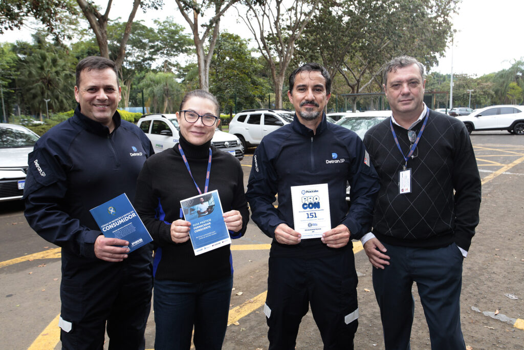 A foto mostra quatro pessoas em pé num estacionamento ao ar livre, sorrindo pra câmera e segurando materiais informativos.Detalhes da cena:Pessoas: Da esquerda pra direita.Homem de uniforme Detran.SP: Usa jaqueta azul-marinho com o logo "Detran.SP" no peito e na manga. Segura o "CÓDIGO DE PROTEÇÃO E DEFESA DO CONSUMIDOR".Mulher: Usa blusa preta de gola alta e óculos. Com um crachá no pescoço, segura o "GUIA PRÁTICO FORNECEDOR CONSCIENTE". Homem de uniforme Detran.SP: Também de jaqueta azul-marinho. Segura um folheto branco do "PROCON Piracicaba 151".Homem de suéter: Usa suéter preto com losangos, camisa social por baixo e crachá com cordão azul. Está com as mãos no bolso.Materiais: Todos os folhetos são da Prefeitura de Piracicaba e tratam de direito do consumidor e fornecimento consciente.Fundo: Estacionamento com vagas demarcadas em amarelo. Tem vários carros brancos ao fundo, árvores e área verde. Parece um espaço institucional ou de órgão público.A imagem passa ideia de ação conjunta entre Detran.SP e Procon Piracicaba, provavelmente em uma campanha ou entrega de materiais educativos ao público.