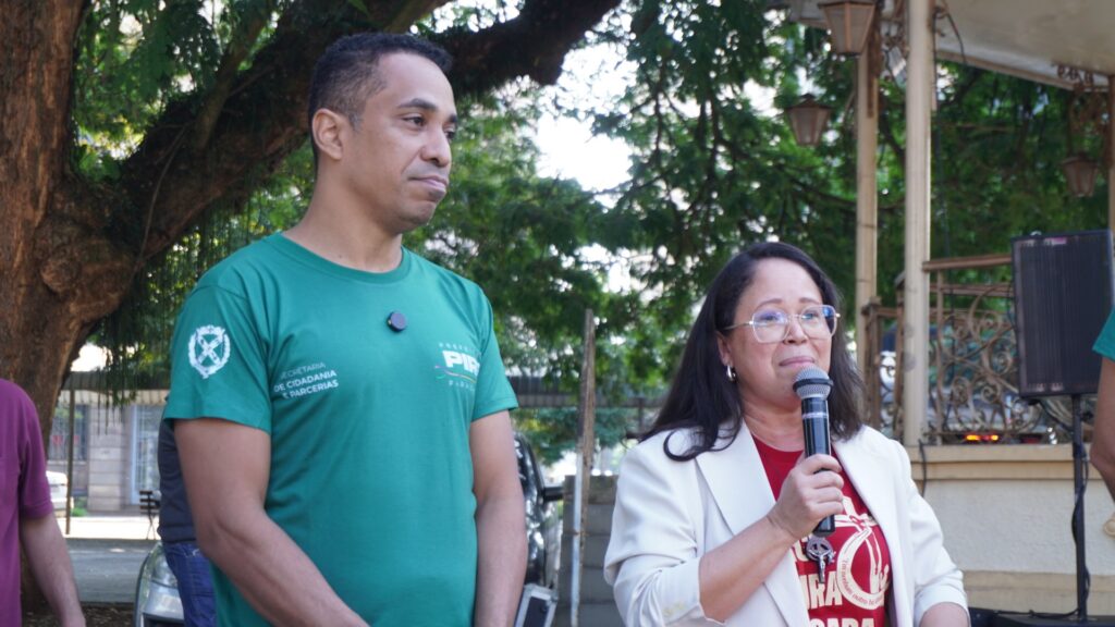 A imagem mostra duas pessoas em um ambiente externo, possivelmente uma praça ou parque, durante um evento ou entrevista. * À esquerda, um homem de pele morena, com cabelos curtos e escuros, veste uma camiseta verde com o logotipo e o texto "Secretaria de Cidadania e Parcerias" da "Prefeitura de Piracicaba. Ele está de pé, com uma expressão séria, olhando para o lado. * À direita, uma mulher de pele clara, com cabelos longos e escuros, usa óculos de grau, um blazer claro sobre uma camiseta vermelha. Ela segura um microfone com a mão direita, parecendo estar falando ou discursando. * O fundo apresenta árvores com folhagem densa, parte de uma estrutura metálica decorativa (semelhante a um coreto) e um alto-falante montado em um suporte. A iluminação parece ser de luz natural do dia.