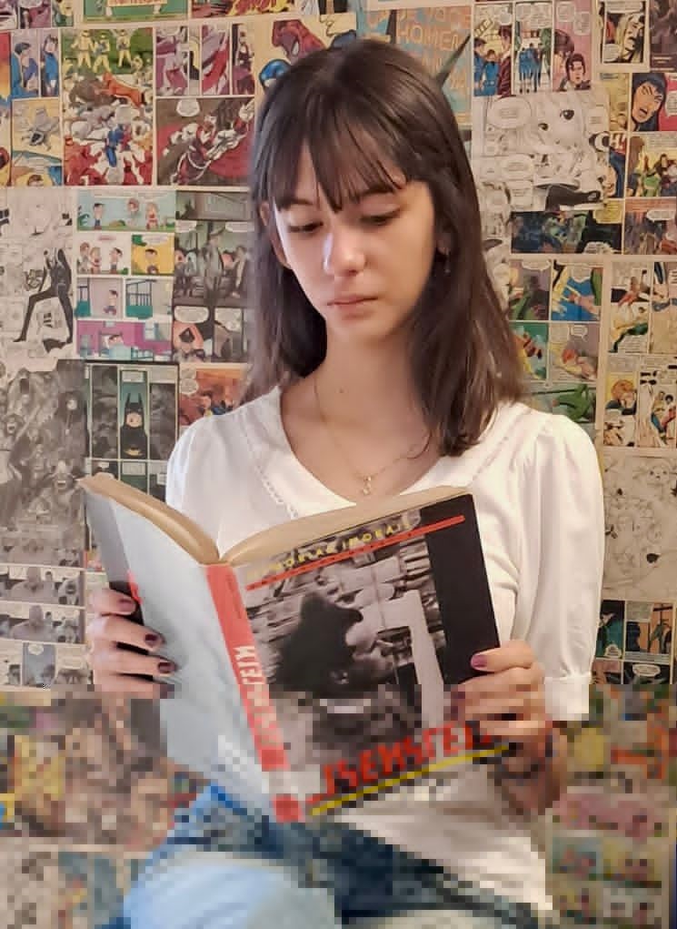 A imagem mostra uma jovem de cabelos castanhos e franja, vestindo uma blusa branca, sentada e concentrada na leitura de um livro. * Ela segura o livro aberto com as duas mãos, e a capa traz uma imagem em preto e branco com o título em letras destacadas. * O fundo da imagem é inteiramente coberto por uma colagem de páginas de histórias em quadrinhos, criando um ambiente temático e vibrante. * A iluminação é suave e o foco principal está no rosto da jovem e no livro que ela lê.