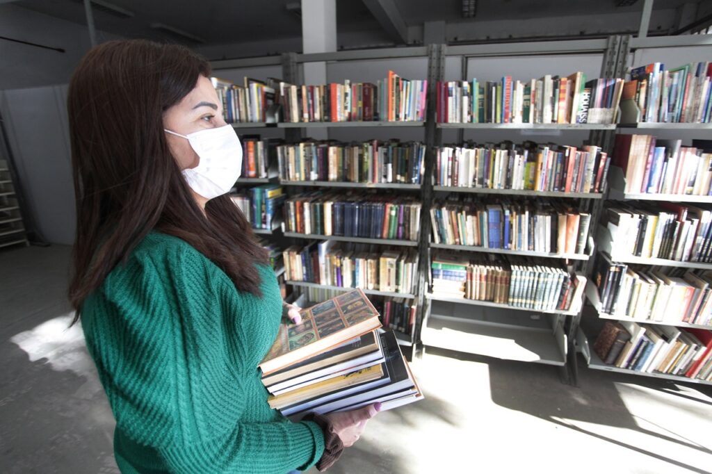 A imagem mostra uma mulher de perfil, voltada para a direita, em um ambiente que parece ser uma biblioteca ou livraria. Ela tem cabelos longos e castanhos escuros, e está usando uma máscara branca de proteção facial. A mulher veste um suéter verde vibrante com textura. Em suas mãos, ela segura uma pilha de quatro livros. Ao fundo, há estantes metálicas preenchidas com muitos livros de diferentes cores e tamanhos. A iluminação é intensa e direcionada, provavelmente vinda de uma janela, criando sombras nítidas no chão. O ambiente é interno, com um visual simples e funcional. A foto foi tirada ao nível dos olhos, com foco claro na mulher e nos livros que ela segura.