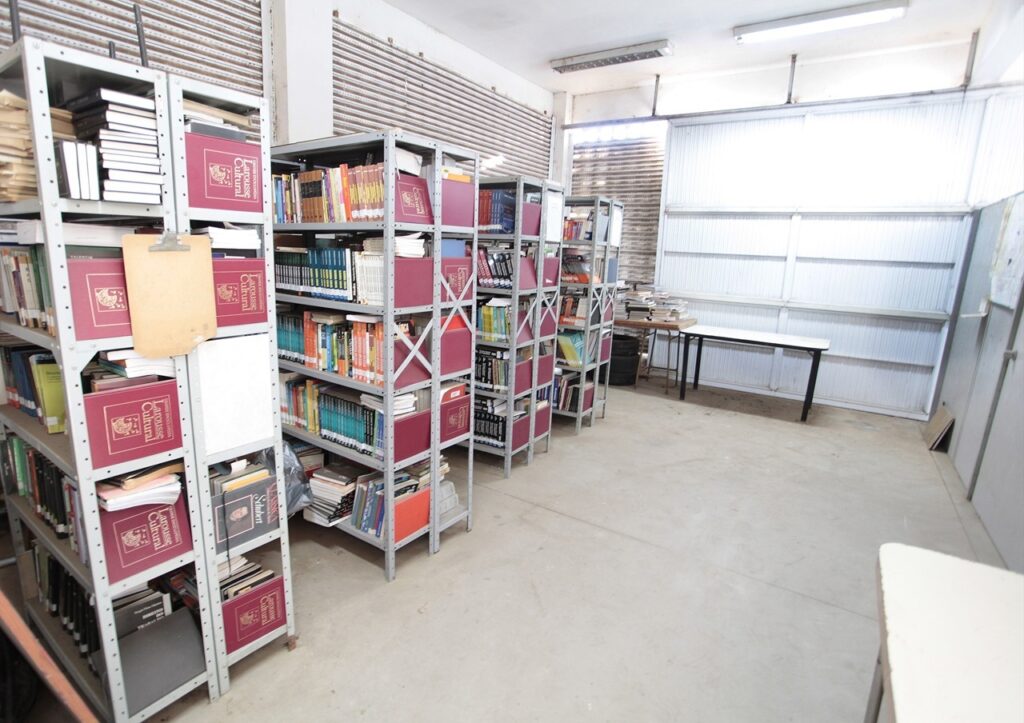 A imagem mostra uma sala de armazenamento ou arquivo, com várias estantes metálicas industriais alinhadas em fileiras paralelas, criando um corredor central. As estantes estão repletas de livros, a maioria com capas vermelhas e com o título "Larousse Cultural" visível em muitos deles. À esquerda, pendurado em uma das estantes, há um quadro de aviso ou prancheta na cor bege. Ao fundo, há uma mesa branca encostada em uma parede feita de painéis metálicos ondulados e janelas grandes, sugerindo um ambiente de galpão ou depósito. O piso é de concreto liso e a iluminação é clara e uniforme, provavelmente proveniente de luzes fluorescentes no teto. O ambiente transmite uma sensação de organização funcional, típica de um arquivo ou biblioteca de armazenamento.