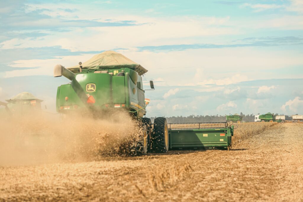 Esta imagem mostra uma cena de colheita em um campo agrícola de grande escala. Destaque: No centro, uma colheitadeira John Deere verde está operando, levantando uma grande nuvem de poeira enquanto corta a vegetação. É possível ver parte de sua plataforma de corte avançando sobre o campo seco. Contexto: O ambiente é um vasto terreno plano e seco, típico de uma lavoura no momento da colheita. Profundidade: Ao fundo, é possível ver outras máquinas agrícolas semelhantes, incluindo uma segunda colheitadeira e caminhões de transporte alinhados, sugerindo uma operação mecanizada em grande escala. Céu: O dia está claro, com um céu azul parcialmente nublado, e a iluminação sugere um momento de trabalho intenso no campo.