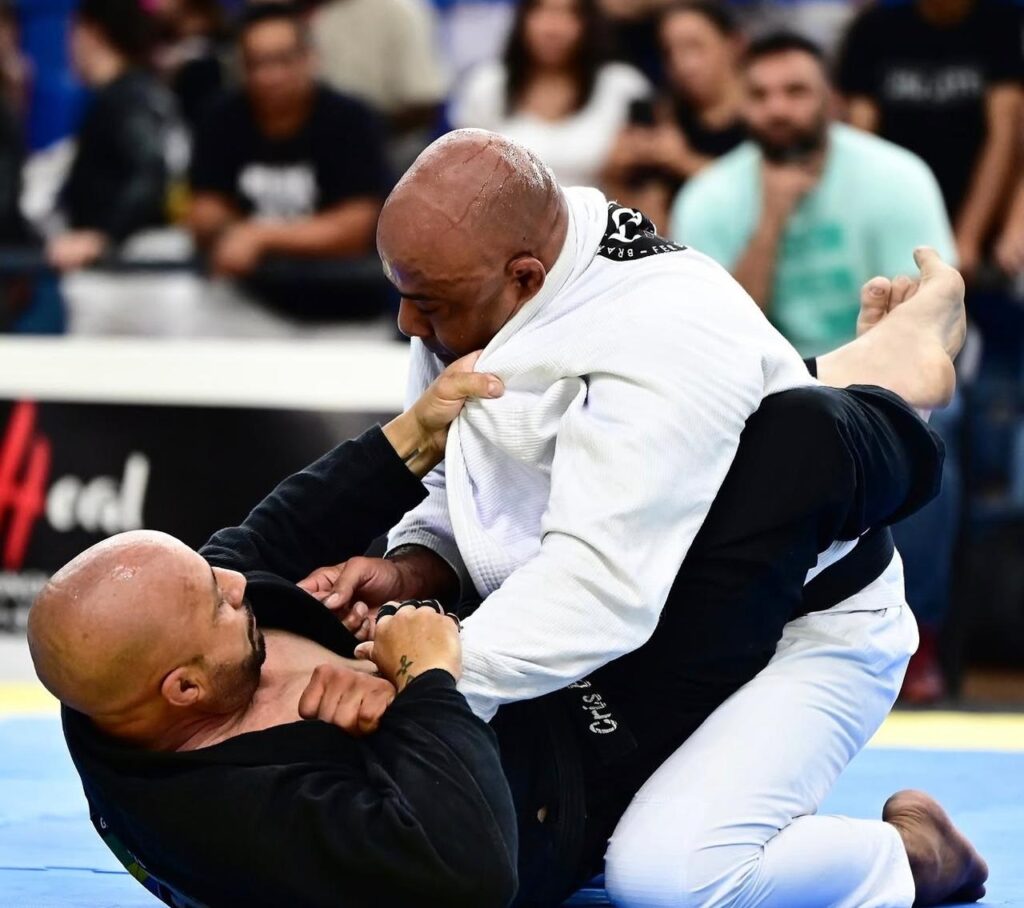 A imagem mostra dois atletas de Jiu-Jitsu em uma disputa intensa durante uma competição. * O atleta de quimono branco está por cima, aplicando uma pressão sobre o adversário, enquanto segura a gola do quimono preto com uma mão e mantém o controle da postura. * O atleta de quimono preto está por baixo, de costas para o tatame, utilizando sua guarda para controlar o oponente e tentar neutralizar a pressão ou buscar uma raspagem. * Ambos os lutadores estão concentrados e suados, evidenciando o esforço físico da luta. Ao fundo, é possível ver parte da plateia acompanhando a disputa, com o ambiente característico de um torneio de artes marciais.