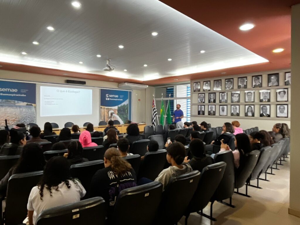 A imagem mostra uma aula ou palestra acontecendo em um auditório ou sala de conferências. Na frente, um homem vestido com uma camiseta azul está de pé, aparentemente explicando algo para o público. Atrás dele, há uma grande tela de projeção que exibe o título "O que é Ecologia?" e alguns pontos em destaque, embora o conteúdo detalhado não seja legível na imagem. O público é composto por várias pessoas sentadas em cadeiras pretas, assistindo à apresentação. Algumas delas estão de costas para a câmera, enquanto outras aparecem de perfil ou olhando para seus celulares. A sala é bem iluminada, com luzes embutidas no teto. Nas paredes laterais, há banners com o logo "semae" e o nome "@semaepiracicaba", indicando que o evento provavelmente está relacionado a essa organização. Também é possível ver duas bandeiras próximas ao palestrante: a bandeira do Brasil e a bandeira do estado de São Paulo. No fundo da sala, uma parede está decorada com uma grade contendo várias fotografias emolduradas, possivelmente retratos de pessoas importantes ou membros da instituição. A atmosfera geral é formal e acadêmica, indicando um ambiente de ensino ou capacitação.