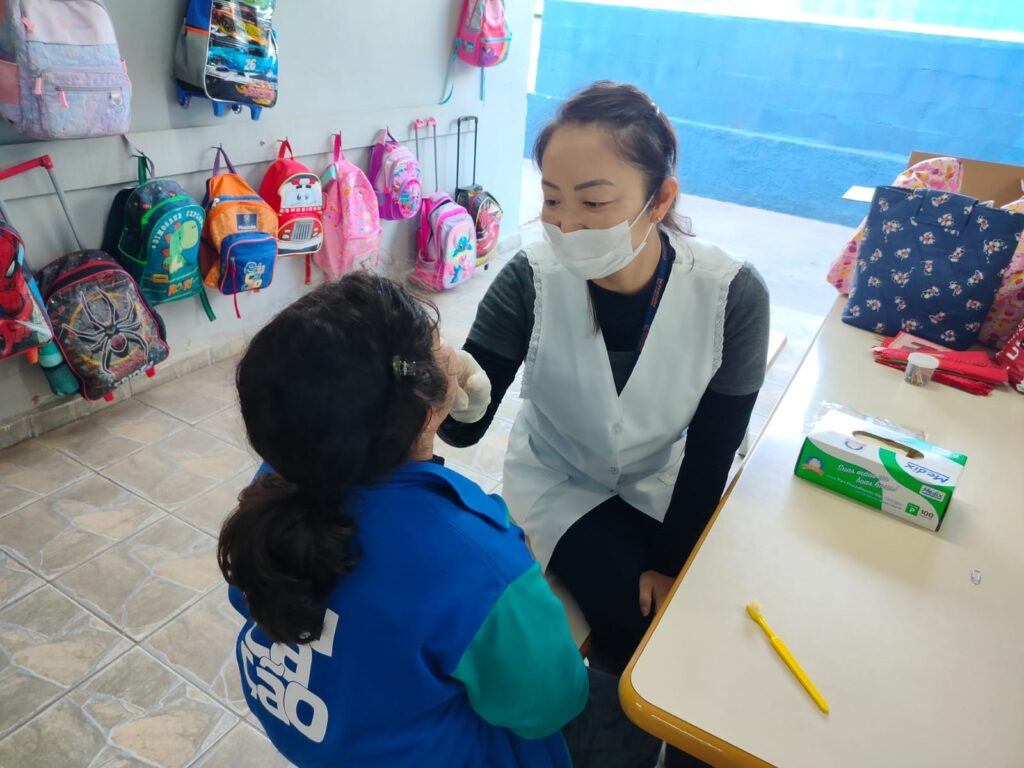 A imagem mostra uma cena de atendimento de saúde ou higiene em um ambiente escolar. * Em primeiro plano, uma profissional de saúde, vestindo um avental branco sobre uma blusa cinza e uma máscara facial, está sentada e examinando a boca de uma criança. Ela utiliza uma luva descartável e sua postura é atenta e profissional. * A criança está de costas para a câmera, vestindo um casaco azul e verde com a inscrição "CRECHE" visível nas costas. Seus cabelos estão presos em um rabo de cavalo. * Sobre a mesa, à direita, há uma caixa de luvas descartáveis da marca Medix e uma escova de dentes amarela. * Ao fundo, a parede está decorada com diversas mochilas escolares coloridas de diferentes tamanhos e estampas, penduradas em ganchos, o que caracteriza o local como uma instituição de ensino infantil. O piso é de cerâmica clara e o ambiente parece bem iluminado.