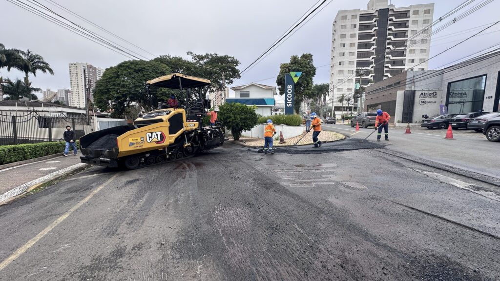 Hoje descreva em um parágrafo Na cena registrada, observa-se uma operação de recapeamento urbano em que um pavimentador Caterpillar deposita nova camada de asfalto enquanto trabalhadores, vestidos com coletes e capacetes laranja, espalham e nivelam o material com ferramentas manuais. O trabalho ocorre em um cruzamento delimitado por cones de sinalização, próximo a prédios comerciais identificados com os nomes Sicoob e BTG_Pactual, compondo um cenário típico de manutenção de infraestrutura viária em área urbana, onde a presença de pedestres e o fluxo cotidiano convivem com a atividade intensa da obra.