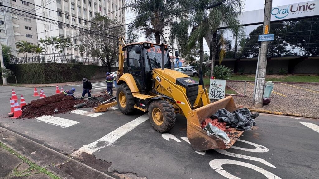 A imagem mostra uma cena urbana durante o dia, onde está acontecendo um trabalho de manutenção ou construção na rua. No centro da imagem, há uma retroescavadeira amarela da marca Caterpillar (modelo 416), posicionada sobre uma faixa de pedestres. Na pá dianteira da máquina, há tijolos vermelhos e sacos plásticos pretos. Três trabalhadores estão agachados perto da traseira da retroescavadeira, aparentemente realizando algum tipo de escavação ou reparo no chão, onde há uma pilha de terra. Ao redor da área de trabalho, estão colocados cones de sinalização laranja para alertar os pedestres e veículos. Ao fundo, vê-se um edifício com várias janelas e algumas árvores, além de uma placa comercial que indica a presença de uma clínica odontológica chamada "Oral Unic Implantes". Também é possível identificar uma placa de rua com o nome "Rua dos Operários". A cena é iluminada por luz natural, em um dia possivelmente nublado.