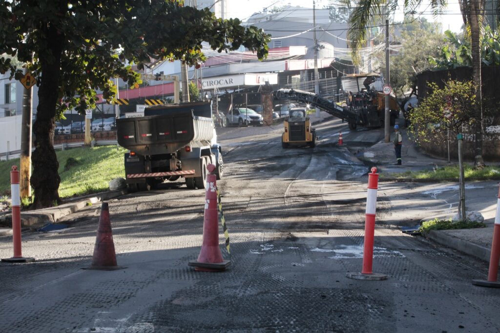 Na imagem, vemos uma cena de obras viárias em área urbana: máquinas pesadas como caminhão basculante, fresadora e carregadeira pequena atuam na manutenção da rua, que apresenta trechos de asfalto removidos. Cones de sinalização delimitam a área de trabalho, enquanto um trabalhador com equipamentos de segurança acompanha a operação ao lado. Ao fundo, aparecem prédios, árvores e uma placa indicando limite de velocidade de 40 km/h. O cenário transmite a importância da infraestrutura urbana e do processo de recapeamento, destacando o papel da segurança no trânsito durante intervenções desse tipo.