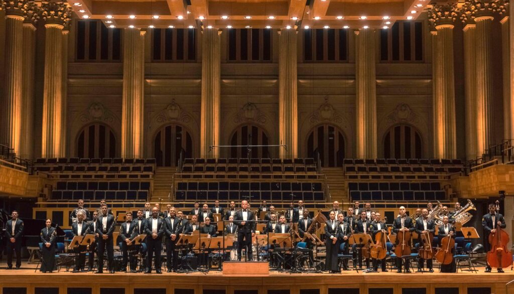 Esta imagem mostra uma orquestra sinfônica completa posicionado no palco de uma sala de concertos imponente e clássica. * Os músicos estão dispostos em semicírculo, vestindo trajes formais pretos, com os instrumentos organizados por naipes, incluindo cordas, sopros e metais. * No centro, um maestro está em um pódio, conduzindo a apresentação, enquanto os instrumentistas estão prontos com seus instrumentos, como violoncelos, contrabaixos, clarinetes e trompas. * O cenário ao fundo é arquitetonicamente rico, caracterizado por colunas coríntias altas, iluminação quente e arcos decorativos, o que confere um tom solene e elegante ao ambiente. * A iluminação do palco é focada nos músicos, criando um contraste com a penumbra das fileiras de assentos vazios da plateia ao fundo, destacando a importância do evento musical.