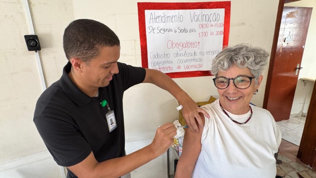 A imagem mostra uma cena de vacinação em um ambiente interno, possivelmente uma clínica ou posto de saúde. À esquerda, um profissional de saúde, vestido com uma camisa preta e crachá, está aplicando uma vacina no braço de uma senhora idosa que está à direita. A senhora tem cabelo grisalho e usa óculos, um colar de contas e uma camiseta branca. Ela sorri durante o momento da aplicação da vacina. Ao fundo, há uma placa branca com borda vermelha brilhante, na qual está escrito em português informações sobre o atendimento de vacinação. O texto indica os horários de atendimento de segunda a sexta-feira, das 08:00 às 15:00 e das 17:00 às 20:00 horas, e lista alguns requisitos obrigatórios para a vacinação, como cadastro atualizado na recepção, apresentação de documentação e a caderneta de vacinação. A iluminação é clara e destaca os dois personagens principais da imagem.