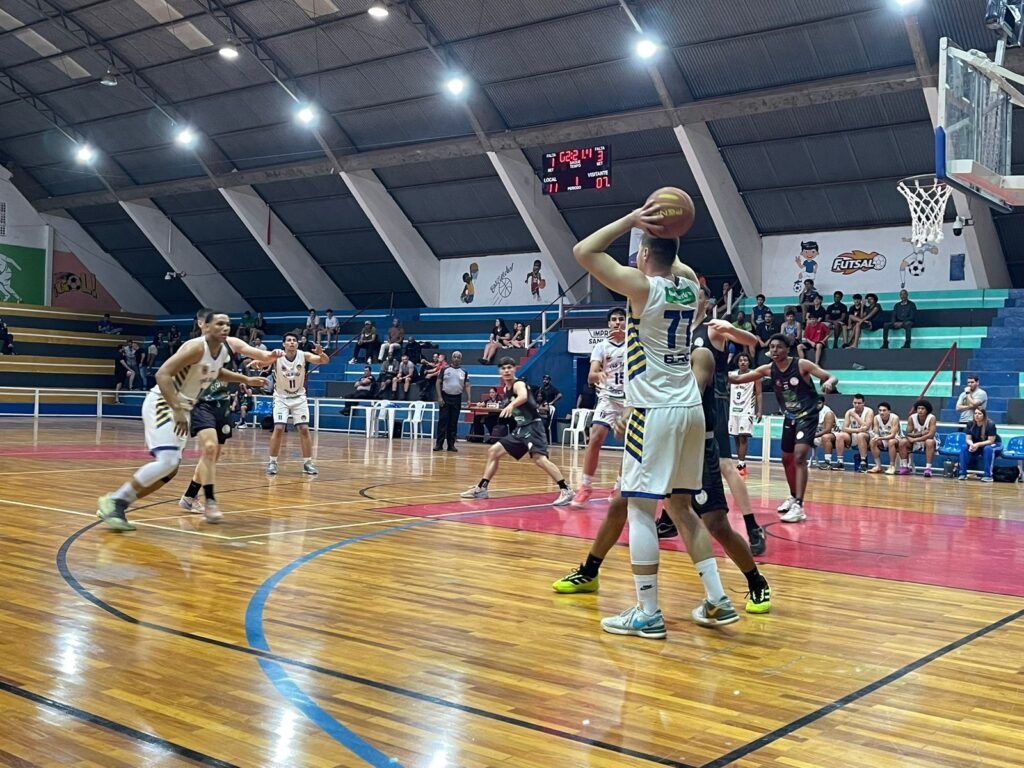 Esta é uma cena dinâmica de uma partida de basquete em uma quadra coberta. Ação principal: Um jogador vestindo uma camisa branca com detalhes azuis e amarelos (número 77) está segurando a bola acima da cabeça, no centro da quadra, aparentemente se preparando para um passe ou arremesso. Contexto: Outros jogadores, vestindo uniformes brancos ou pretos, estão posicionados ao redor dele, atentos à jogada. Ambiente: Ao fundo, há um placar eletrônico indicando "11" para o time local e "07" para o visitante, com uma arquibancada ocupada por espectadores assistindo ao jogo.