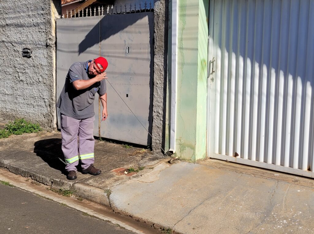 Esta é uma cena cotidiana de trabalho de manutenção urbana. Ação: Um funcionário, usando um uniforme cinza com faixas refletivas na calça e um boné vermelho, está inclinado sobre a calçada. Ele segura uma haste metálica que toca um ponto no chão perto de uma descida de calha (onde parece haver um ralo ou acesso à rede), enquanto leva a mão ao rosto, parecendo concentrado ou ajustando algo. Ambiente: A cena acontece em frente a uma residência com um portão de garagem branco e uma parede verde-clara. O muro à esquerda tem um acabamento texturizado. Detalhes: O dia parece ensolarado, projetando sombras nítidas no portão e no chão. O trabalhador usa botas de segurança pretas e o uniforme sugere que ele está em serviço profissional de inspeção ou reparo.