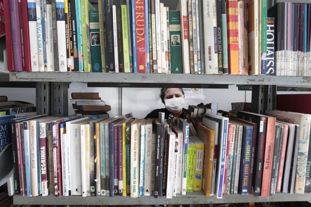 A imagem mostra duas estantes cheias de livros, organizados verticalmente, com diversas cores e títulos visíveis nas lombadas. Entre as estantes, aparece uma pessoa usando máscara branca, olhando diretamente para a câmera. A pessoa está enquadrada no centro da imagem, com os livros acima e abaixo dela formando um cenário ao redor. Os títulos dos livros são variados, incluindo autores brasileiros renomados como Jorge Amado, Vinicius de Moraes, e Monteiro Lobato, além de nomes internacionais como Kurt Vonnegut e Pablo Neruda. O ambiente parece ser uma biblioteca ou livraria, com iluminação interna e um clima de silêncio e concentração. A máscara indica que a foto foi provavelmente tirada durante a pandemia de COVID-19. A imagem transmite uma sensação de imersão no mundo literário, com a figura humana destacada em meio ao grande volume de livros.