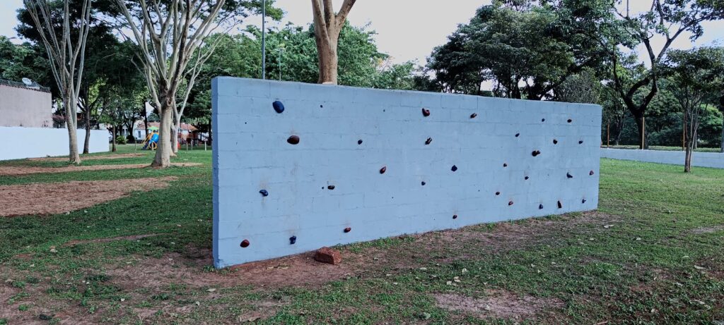 A imagem mostra uma parede de escalada ao ar livre, feita de concreto pintado de azul claro. A parede é retangular, posicionada horizontalmente sobre uma área gramada em um parque. Nela estão fixas várias agarras coloridas (vermelhas, azuis e pretas), usadas para escalada. Ao fundo, vê-se uma área verde com árvores e um playground com estruturas em azul e amarelo. O chão ao redor da parede é uma mistura de grama e terra. A cena está iluminada pela luz natural do dia, e não há pessoas presentes na imagem.