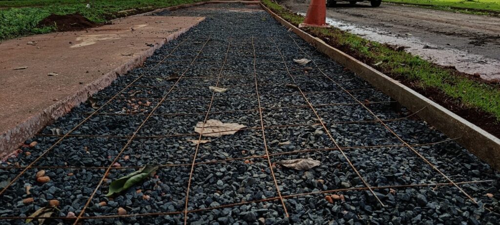 A imagem mostra uma obra de construção civil em andamento, focada na preparação de uma calçada ou caminho. * O elemento central é uma camada de brita (pedras pequenas) que serve como base para o pavimento. Sobre essa base, foi estendida uma malha de aço (tela soldada), que servirá para estruturar e reforçar o concreto que será aplicado posteriormente. * À esquerda, é possível ver um trecho de calçada já concretado, indicando que o trabalho é uma extensão ou reparo. A área está delimitada por formas de madeira nas laterais, que servem como guia para o nivelamento do concreto. * O ambiente ao redor sugere uma área externa urbana ou suburbana, com gramado, terra e uma rua de terra batida ao fundo, onde um cone de trânsito está posicionado. A iluminação indica um dia nublado ou final de tarde.