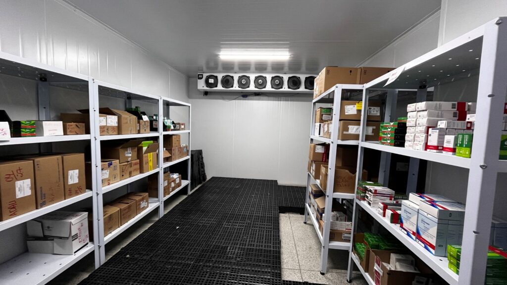 A imagem mostra o interior de uma câmara frigorífica ou sala de armazenamento refrigerado, provavelmente utilizada para guardar medicamentos ou produtos farmacêuticos que precisam ser mantidos em baixas temperaturas. O ambiente é limpo e organizado, com duas fileiras de estantes metálicas brancas alinhadas paralelamente nas laterais do espaço. As estantes estão cheias de caixas de papelão e embalagens diversas, muitas delas com etiquetas manuscritas ou impressas, indicando o conteúdo ou datas. O piso é coberto por um tipo de grade preta plástica, provavelmente para facilitar a ventilação e drenagem. Na parede do fundo, há uma grande unidade de refrigeração com múltiplas ventoinhas visíveis, responsável por manter a temperatura controlada no ambiente. A iluminação é feita por uma luminária fluorescente instalada no teto, que proporciona uma luz branca e uniforme em todo o espaço. No geral, a imagem transmite a ideia de um local controlado e organizado, essencial para a conservação adequada de insumos sensíveis, como medicamentos ou vacinas, garantindo a sua integridade e eficácia.