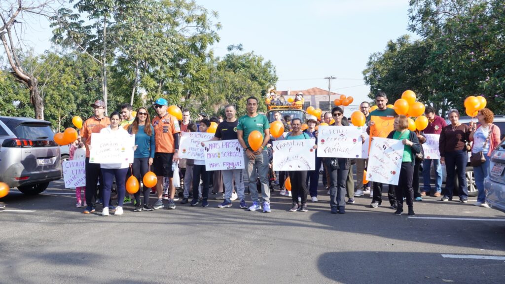 A imagem retrata um grupo de pessoas de diversas idades participando de uma caminhada ou evento comunitário ao ar livre, possivelmente relacionado à valorização da família. * O grupo está posicionado em uma rua asfaltada, segurando cartazes com mensagens positivas e informativas sobre a importância da família, como "Mais passos, mais sorrisos, mais família!" e "O futuro começa na família". * Muitas pessoas seguram balões cor de laranja, que parecem ser a cor temática do evento. O ambiente é ensolarado e arborizado, com carros estacionados nas laterais da via. * Os participantes vestem roupas casuais e esportivas, e alguns vestem camisetas padronizadas na cor verde, sugerindo que fazem parte de um mesmo grupo, organização ou projeto social. A atmosfera geral é de união, tranquilidade e engajamento comunitário.