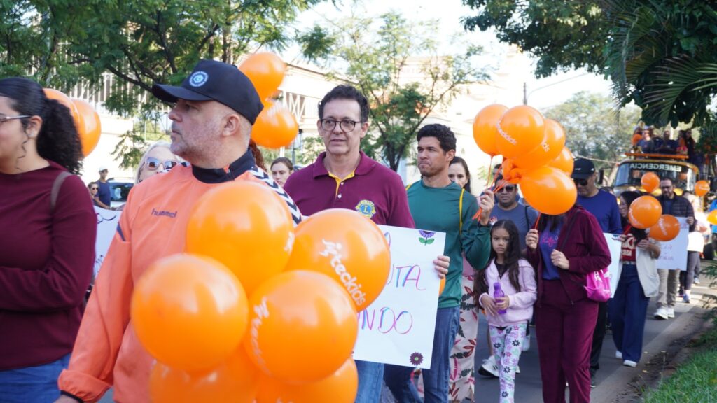 A imagem retrata um grupo de pessoas caminhando ao ar livre durante o que parece ser uma marcha ou evento de conscientização. * Em primeiro plano, um homem de boné preto e jaqueta laranja segura um grande cacho de balões cor de laranja. Ao lado dele, um homem de óculos veste uma camisa polo bordô com o emblema do Lions Club. * Várias pessoas ao redor carregam cartazes com mensagens escritas à mão e balões laranjas, que possuem a marca "inteckids" estampada. * O ambiente é iluminado pela luz do dia, com árvores ao fundo e um veículo de apoio, possivelmente um caminhão de som, visível mais à frente. As pessoas caminham por uma via pavimentada, sugerindo um evento comunitário ou uma passeata em um espaço público.