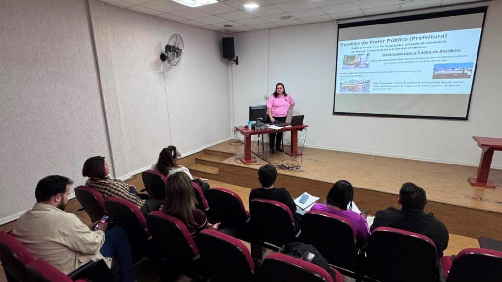 A foto mostra uma palestra ou treinamento acontecendo num pequeno auditório.Cena principal:Palestrante: Uma mulher de camiseta rosa está de pé atrás de uma mesa no palco. Ela parece conduzir a apresentação.Slide projetado: O tema é "Deveres do Poder Público (Prefeitura)" e fala sobre a Prefeitura de Piracicaba. O slide detalha o "Gerenciamento e Coleta de Resíduos", incluindo coleta de lixo domiciliar, coleta seletiva e "Cata Cacareco" para móveis e utensílios em desuso.Público: Sete pessoas estão sentadas nas cadeiras vermelhas do auditório, de costas pra câmera. Algumas olham pra frente, outras mexem no celular ou anotam. São as mesmas pessoas da foto anterior com as placas de "MITO ou VERDADE".Ambiente: Sala com piso e palco de madeira, paredes claras, ventilador na parede e caixas de som. Na mesa da palestrante tem notebook, monitor e papéis.O contexto é uma capacitação sobre gestão de resíduos e deveres da prefeitura, provavelmente parte do mesmo evento da dinâmica de "Mito ou Verdade".