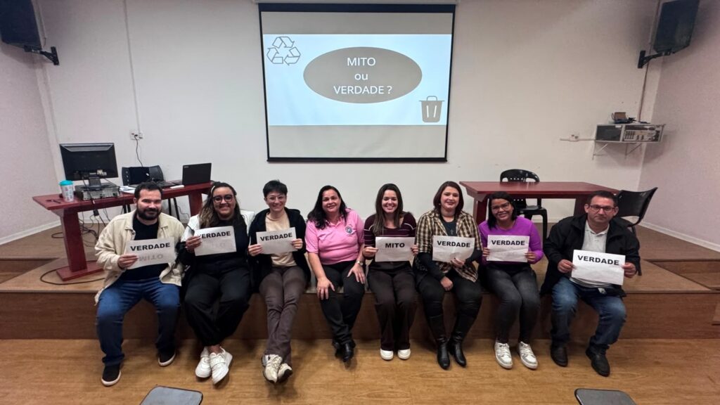 A imagem mostra um grupo de 8 pessoas sentadas lado a lado num pequeno palco ou degrau de madeira, numa sala que parece ser de aula ou treinamento.Detalhes da cena:Ao fundo: Um projetor exibe o slide "MITO ou VERDADE?" com ícones de reciclagem e de uma lixeira.Pessoas: Todos estão sorrindo pra foto. Sete pessoas seguram placas com a palavra "VERDADE" e uma pessoa segura uma placa com "MITO".Ambiente: A sala tem paredes brancas, piso de madeira e mesas com computadores e notebooks nos cantos. Parece um espaço de capacitação ou workshop.Vestimenta: Roupas casuais. Tem camiseta rosa, camisa xadrez, moletom roxo, jaqueta preta. Estilo bem diverso.O contexto geral é de uma dinâmica ou atividade educativa, provavelmente sobre reciclagem ou meio ambiente, onde os participantes respondem se uma afirmação é mito ou verdade.