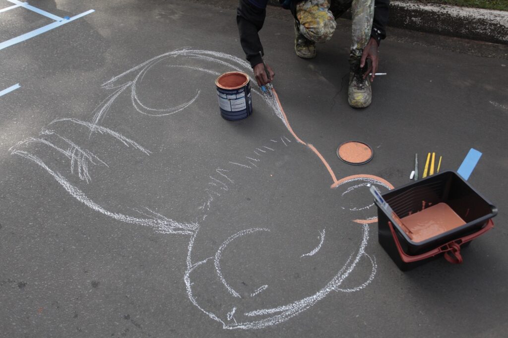 A imagem mostra um artista pintando um mural diretamente no asfalto escuro de uma rua ou estacionamento. A pessoa está agachada, usando calças de trabalho manchadas de tinta, uma blusa preta de manga longa e botas desgastadas. O artista está contornando o desenho de um animal — possivelmente uma capivara que está esboçado com giz branco no chão. O desenho está parcialmente pintado com tinta de cor pêssego, que está em uma bandeja plástica preta ao lado do artista, junto com pincéis. Também há uma lata de tinta da marca "Coral" aberta próxima ao artista. O ambiente parece ser ao ar livre, com luz natural e linhas azuis pintadas no chão ao fundo. A imagem destaca o processo criativo e o contraste entre a textura áspera do asfalto e as cores suaves da pintura.