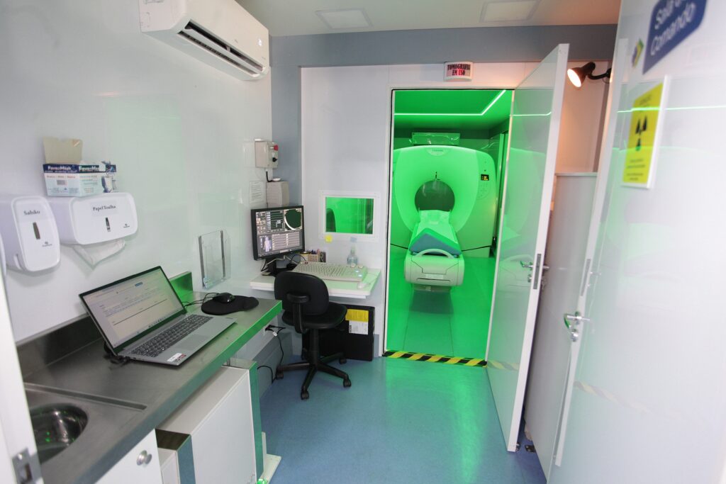 Esta imagem mostra a sala de controle e a sala de exames de uma unidade móvel de tomografia computadorizada. • Sala de Controle: Em primeiro plano, há uma bancada com um notebook aberto, um computador de mesa com monitor e teclado, e dispensadores de sabão e papel toalha. O ambiente é limpo e funcional. • Sala de Exames: Através de uma porta aberta, vê-se a sala onde o equipamento de tomografia está instalado. O espaço está iluminado por uma luz verde intensa. • Equipamento: O tomógrafo, da marca Toshiba, está posicionado no centro da sala ao fundo. Acima da porta, há uma placa indicando "TOMOGRAFIA EM USO". • Sinalização: Na porta lateral direita, há um aviso de radiação ionizante (o símbolo de perigo amarelo e preto), indicando que se trata de uma área de controle radiológico.