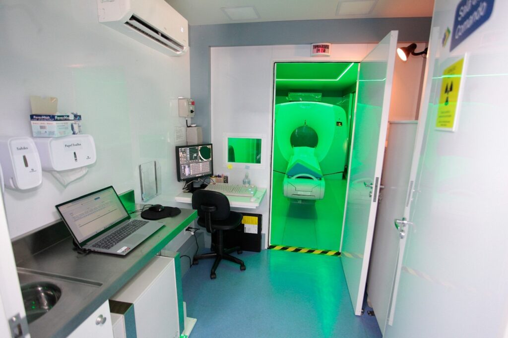 Esta imagem mostra o interior de uma unidade móvel de saúde, especificamente uma sala de comando para exames de tomografia computadorizada. * No lado esquerdo, há uma bancada de aço inoxidável com um notebook aberto, um dispensador de sabonete e um suporte para papel toalha. Acima, um aparelho de ar-condicionado está fixado na parede. * No centro, encontra-se uma estação de trabalho com um monitor, teclado e uma cadeira giratória preta. Uma janela de vidro permite a visualização direta da sala do equipamento. * À direita, uma porta aberta revela a sala do tomógrafo, que possui uma iluminação verde intensa. O equipamento de tomografia, da marca Toshiba, está posicionado ao centro, pronto para uso, com uma placa acima da porta indicando "TOMOGRAFIA EM USO". * O ambiente é funcional e compacto, com piso azul e paredes brancas, seguindo padrões de higiene e sinalização de segurança radiológica.