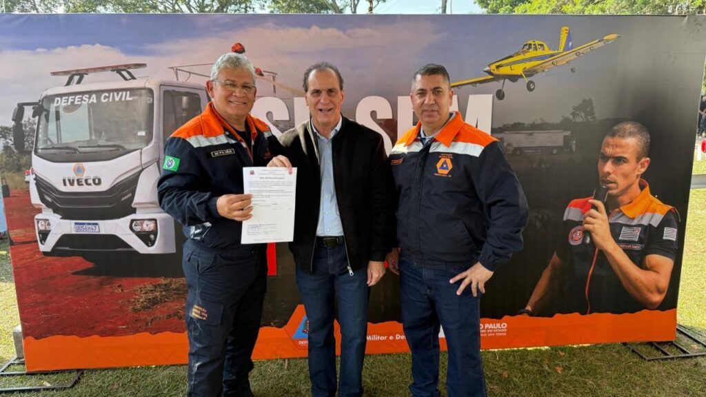 A imagem mostra três homens em pé, posando para uma foto em um evento oficial. Dois deles estão vestidos com uniformes da Defesa Civil, nas cores azul e laranja, enquanto o homem do meio veste uma jaqueta escura e calça jeans. O homem à esquerda segura um documento branco. Atrás deles, há um grande banner promocional que exibe imagens relacionadas à Defesa Civil, incluindo um caminhão Iveco, um avião amarelo de combate a incêndios e uma figura de uma pessoa falando ao microfone. No banner, aparecem textos como "DEFESA CIVIL", "SÃO PAULO", "GOVERNO DO ESTADO" e "PIRACICABA - SP". A cena parece ser uma cerimônia ou evento oficial de entrega ou apresentação de equipamentos para a Defesa Civil do estado de São Paulo. A iluminação é natural, indicando que a foto foi tirada durante o dia.
