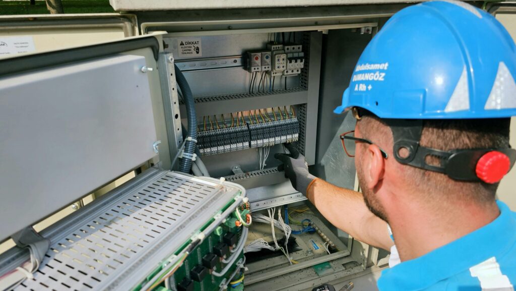 A imagem mostra um técnico eletricista em um ambiente externo, realizando a manutenção ou inspeção em um painel elétrico industrial. * O profissional está usando um capacete de segurança azul, óculos de proteção e luvas, demonstrando o cumprimento de normas de segurança do trabalho. * Ele está com a mão direita dentro do painel, manipulando ou verificando uma régua de bornes (blocos de terminais), onde diversos fios elétricos estão conectados de forma organizada. * O interior do painel contém componentes típicos de automação e controle, como disjuntores, bornes de conexão e uma placa de circuito eletrônico visível na parte inferior esquerda. * Há uma placa de advertência na parte superior interna do painel com o texto em turco "DİKKAT ELEKTRİK ÇARPMA TEHLİKESİ", que significa "Cuidado, perigo de choque elétrico".