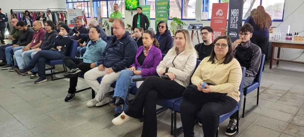 A imagem mostra um grupo de pessoas sentadas em cadeiras azuis, dispostas em fileiras, aparentemente participando de uma palestra, curso ou seminário em um ambiente interno amplo e bem iluminado. O espaço tem piso de azulejos claros e paredes claras, com janelas grandes ao fundo que deixam entrar bastante luz natural. As pessoas são de diferentes idades e gêneros, vestindo roupas casuais e sociais, como blazers, suéteres e camisas. Algumas estão olhando para frente, atentas ao que parece ser um palestrante ou apresentação fora do quadro, enquanto outras olham para a câmera. Ao fundo, há banners e painéis com textos que indicam instituições e temas relacionados à educação e energia, como "SENAI", "Escola SENAI 'Mário Dedini'", "DESCONTO NA FATURA DE ENERGIA SEM INVESTIMENTO", "MBA USP ESALQ" e "SOMOS AGENTES DA MUDANÇA". Também é possível ver um cabideiro com roupas penduradas. O ambiente sugere um evento educacional ou corporativo, voltado para capacitação, inovação ou sustentabilidade.