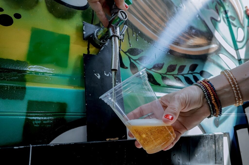 A imagem mostra uma cena próxima e em nível dos olhos, onde uma cerveja está sendo servida de uma torneira para um copo plástico transparente. A mão que segura o copo está adornada com várias pulseiras coloridas de contas e tem as unhas pintadas de vermelho vibrante. O líquido dourado da cerveja está sendo despejado, formando uma pequena camada de espuma branca no topo. Ao fundo, há um mural colorido com grafite em tons de verde, amarelo e preto, com desenhos que lembram folhas estilizadas. A iluminação é natural e destaca a transparência do copo e a cor da cerveja, transmitindo uma atmosfera descontraída e festiva.