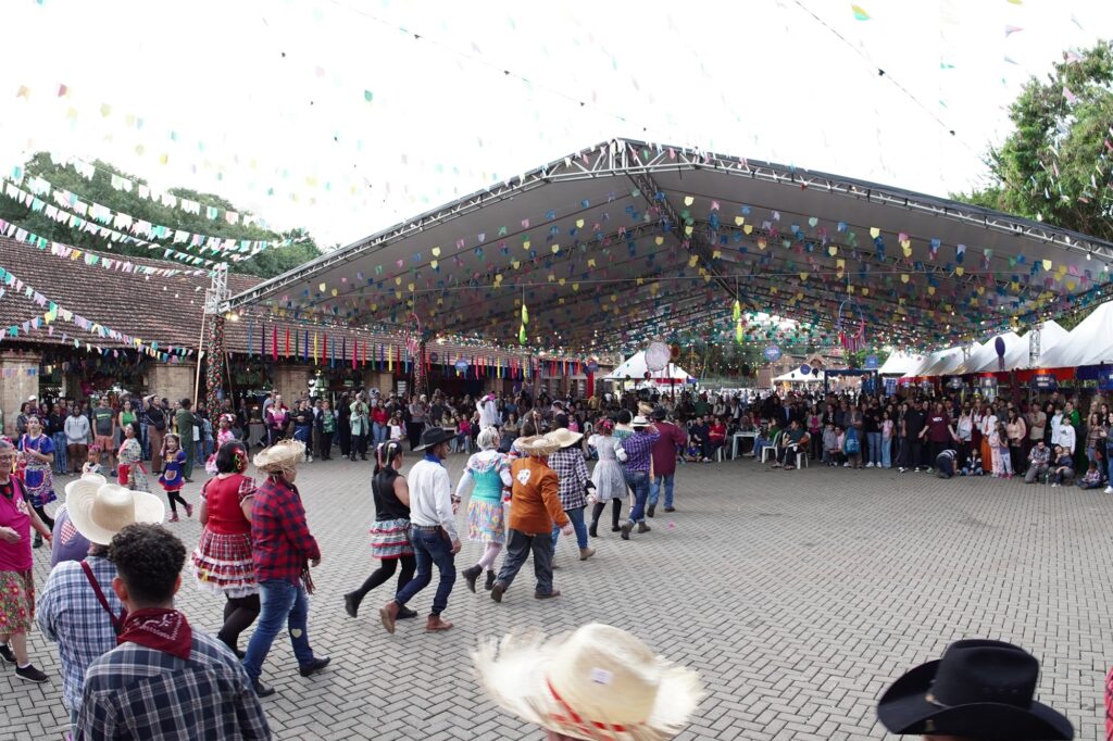 A imagem mostra uma festa junina ao ar livre, com várias pessoas dançando em fila em uma grande praça pavimentada. Os dançarinos estão vestidos com roupas típicas da festa, como camisas xadrez, chapéus de palha e saias coloridas. Acima deles, há bandeirinhas coloridas penduradas, que são tradicionais nas festas juninas. O local é coberto por uma estrutura metálica moderna, que oferece sombra, e ao fundo há uma multidão de pessoas observando a dança, além de algumas barracas brancas e edifícios de tijolos aparentes. A cena transmite um clima alegre e festivo, típico das celebrações culturais brasileiras realizadas no mês de junho.