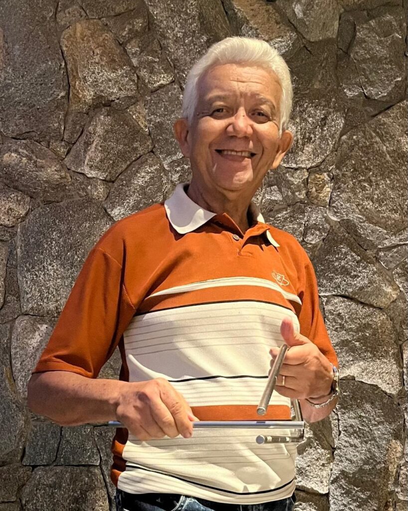A imagem mostra um homem idoso, com cabelos brancos, sorrindo de forma amigável para a câmera. Ele veste uma camisa polo listrada nas cores laranja queimado, branco e preto, com gola branca. Ele segura um instrumento musical de metal, conhecido como triângulo, com uma mão segurando a armação e a outra segurando a baqueta para tocar. No pulso esquerdo, ele usa um relógio metálico. O fundo é uma parede rústica feita de pedras naturais de diferentes tons de cinza e marrom, que adiciona textura e profundidade à imagem. A iluminação é suave, destacando tanto o rosto do homem quanto a textura das pedras ao fundo. A foto está enquadrada em plano médio, focando no homem e no triângulo, transmitindo uma sensação de serenidade e alegria.