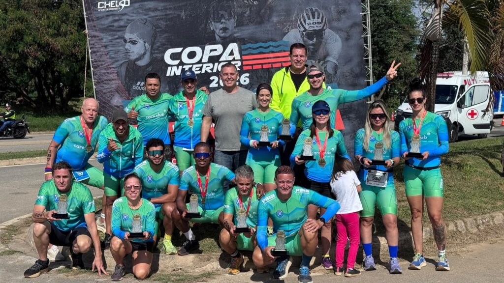 A imagem mostra um grupo de aproximadamente 16 pessoas posando para uma foto em grupo após um evento esportivo, possivelmente uma competição de triatlo ou ciclismo, realizada em 2026, conforme indicado pelo banner ao fundo que diz "COPA INTERIOR 2026". A maioria dos participantes veste uniformes esportivos combinando nas cores verde e azul, com a marca "care club" estampada. Alguns estão segurando troféus pequenos, sugerindo que houve uma premiação. Também há uma criança pequena, vestida de branco e rosa, próxima ao centro do grupo. O ambiente é ao ar livre, com árvores ao fundo e um veículo de ambulância visível à direita, indicando preocupação com a segurança do evento. Todos parecem felizes e comemorativos, com sorrisos e posturas descontraídas, organizados em duas fileiras, alguns agachados na frente e outros em pé atrás. Ao fundo, há um grande banner com os textos "CHELSO SPORTS & BUSINESS" e "COPA INTERIOR 2026", além de números que provavelmente se referem a identificação de atletas ou equipes. A foto foi tirada durante o dia, com boa iluminação natural.