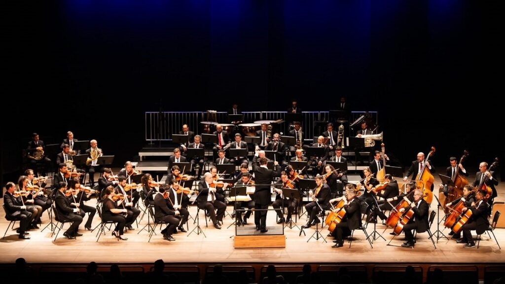 A imagem mostra uma orquestra sinfônica completa, vista de frente e em ângulo aberto. No centro, há um maestro de pé em um pequeno púlpito, de costas para a câmera, conduzindo os músicos. A orquestra está organizada de forma tradicional, com os instrumentos de cordas na frente, as madeiras e metais no meio, e a percussão ao fundo, em plataformas elevadas. Todos os músicos estão vestidos de preto, com trajes formais, e parecem concentrados na execução da música. A iluminação destaca o palco de madeira, criando um ambiente acolhedor e profissional, enquanto o fundo é de um azul escuro que se transforma em preto no topo, dando profundidade à cena. A imagem transmite a energia e a seriedade de uma apresentação musical ao vivo.