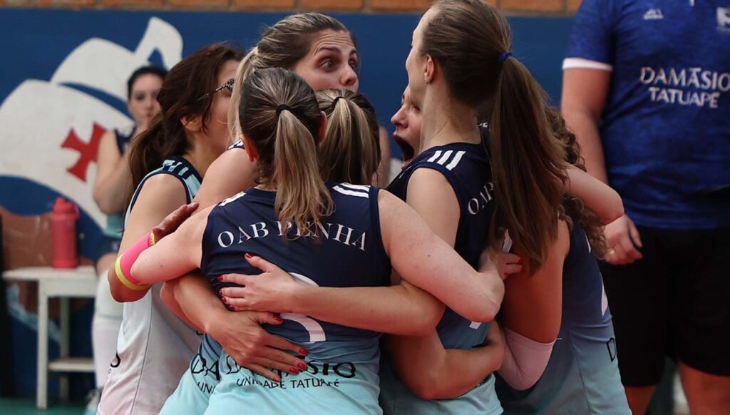 A imagem mostra um grupo de jogadoras de vôlei feminino reunidas em um abraço coletivo, celebrando um momento de euforia e vitória durante uma partida. * Elas vestem uniformes nas cores azul-marinho e azul-claro, com a inscrição "OAB PENHA" visível nas costas de algumas jogadoras. * As expressões faciais demonstram grande intensidade, com algumas jogadoras gritando ou rindo, destacando o espírito de equipe e a emoção do esporte. * Ao fundo, é possível ver parte de uma quadra, com uma parede que exibe o brasão da Ordem dos Advogados do Brasil e uma pessoa à direita vestindo uma camisa com a inscrição "DAMÁSIO TATUAPÉ".