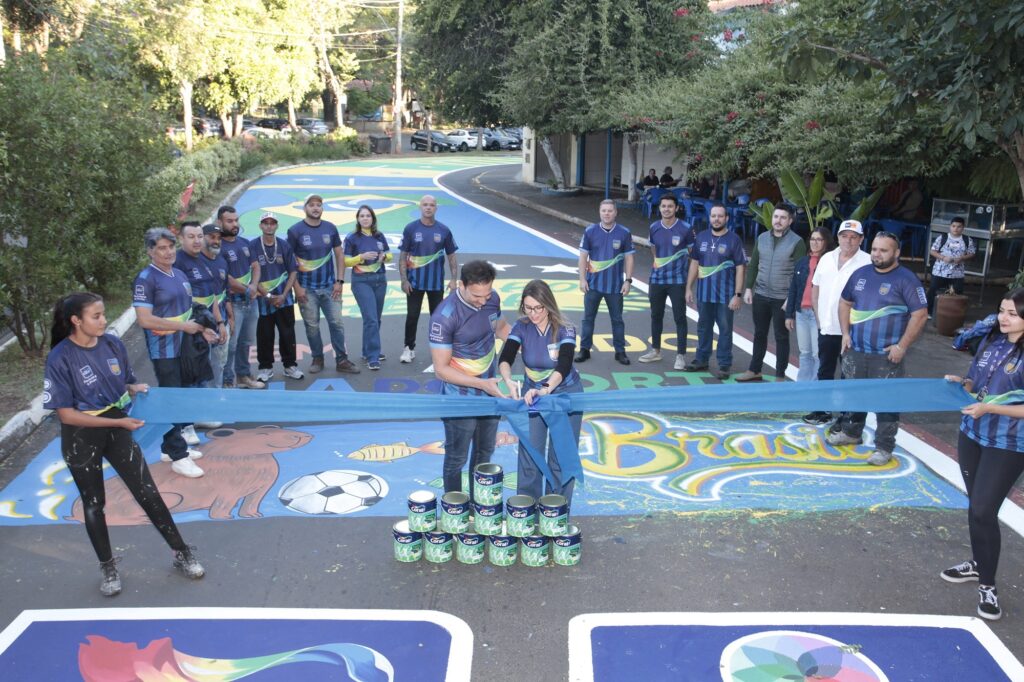 A imagem mostra uma cerimônia de inauguração ao ar livre, provavelmente de uma pintura ou revitalização urbana. No centro, um homem e uma mulher estão cortando uma fita azul, simbolizando a abertura oficial do evento. Ambos vestem camisas azuis com detalhes coloridos, possivelmente uniformes da empresa Coral, que é a marca visível nas latas de tinta empilhadas à frente deles. Ao redor do casal principal, há um grupo de pessoas, a maioria homens, também usando camisas azuis similares, sugerindo que fazem parte da mesma equipe ou organização. Duas mulheres seguram as extremidades da fita, uma de cada lado. O chão está decorado com pinturas coloridas que incluem a bandeira do Brasil, um capivara, uma bola de futebol e a palavra "Brasil", indicando que o evento tem temática nacional e celebra a cultura brasileira. O ambiente é ao ar livre, com árvores e edifícios ao fundo, em um dia claro. A atmosfera é de celebração e formalidade, marcando o início de um projeto ou iniciativa ligada à pintura urbana, com a presença da marca Coral evidenciada pelas latas de tinta e os uniformes.