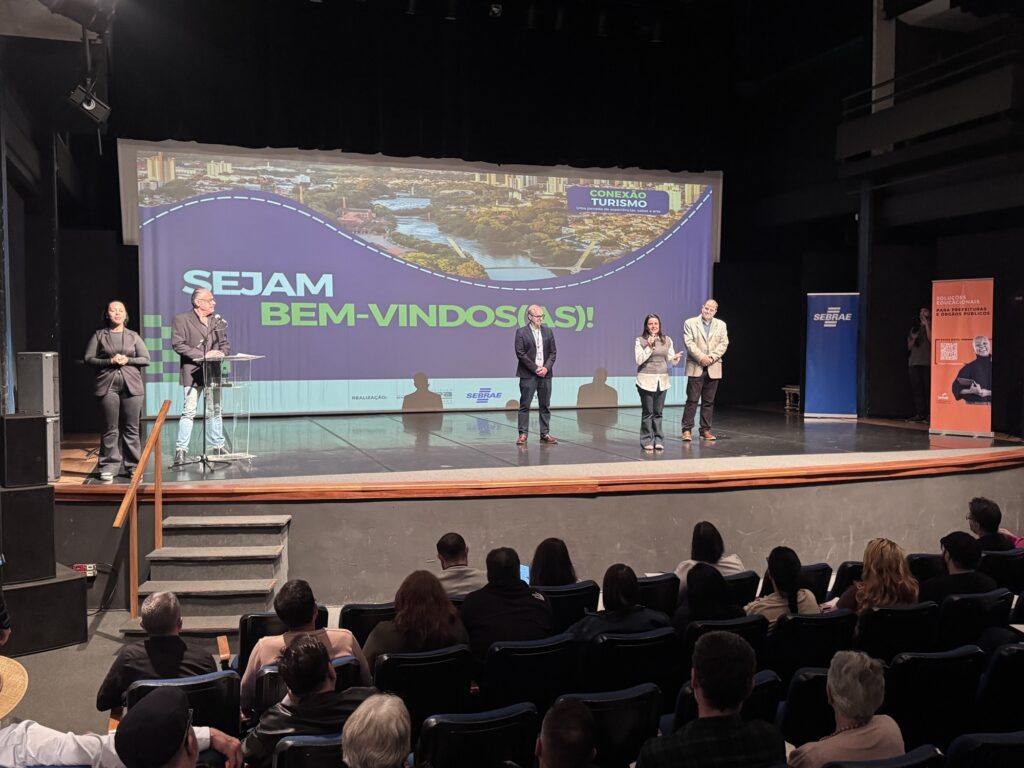 Esta imagem mostra um evento em um auditório, com cinco pessoas no palco e uma plateia sentada. No telão ao fundo, lê-se "SEJAM BEM-VINDOS(AS!)" com o título "CONEXÃO TURISMO", além de uma imagem aérea de uma cidade. O logo do SEBRAE aparece tanto na projeção quanto em um banner vertical à direita. No palco, vemos quatro pessoas: À esquerda, uma mulher de preto em pé. Ao lado dela, um homem de blazer escuro em um púlpito com microfone. Mais ao centro, um homem de camisa azul e blazer, e uma mulher falando ao microfone, vestindo uma blusa branca e colete cinza. À direita, um homem de blazer claro. A plateia está sentada em poltronas escuras, observando o palco. O ambiente parece ser um teatro ou auditório bem iluminado.