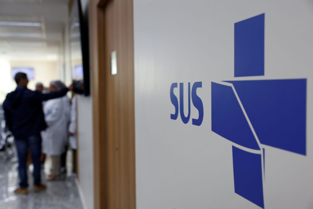 A imagem mostra um ambiente interno de uma unidade de saúde, possivelmente um hospital ou posto de saúde, com foco em uma parede branca onde está estampado o logotipo do SUS (Sistema Único de Saúde) em azul. O logotipo é composto por uma cruz estilizada e a sigla "SUS" ao lado. Ao fundo, desfocado, é possível ver um corredor com algumas pessoas, incluindo profissionais de saúde usando jalecos brancos, o que reforça o contexto médico. A iluminação é clara e o ambiente parece limpo e organizado. A imagem transmite a ideia de um espaço dedicado ao atendimento público de saúde no Brasil.