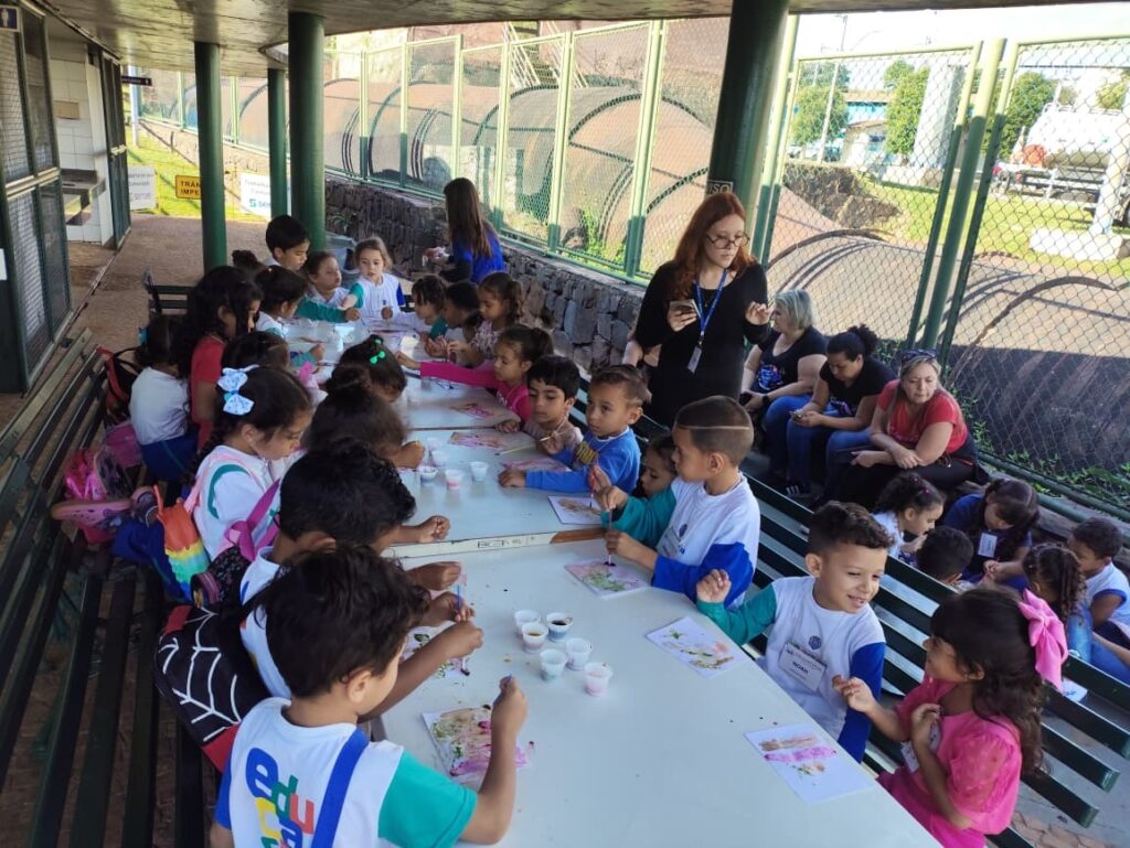 A imagem mostra um grupo de crianças pequenas participando de uma atividade artística ao ar livre, sob uma cobertura que parece ser de uma escola ou centro comunitário. Elas estão sentadas em longas mesas, pintando em pequenas folhas de papel. As crianças estão concentradas em suas pinturas, e várias delas usam uniformes escolares. Há alguns adultos presentes, provavelmente professores ou monitores, que acompanham e auxiliam as crianças. O ambiente é iluminado pela luz natural do dia e ao fundo há uma cerca de arame e uma parede de pedra. O cenário transmite uma atmosfera educativa e de aprendizado coletivo, com as crianças engajadas e participativas na atividade.