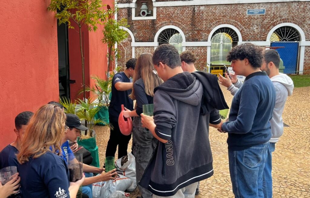 A imagem mostra um grupo de jovens, possivelmente estudantes, participando de uma atividade ao ar livre relacionada ao plantio ou jardinagem. Eles estão reunidos em um espaço aberto, provavelmente um pátio, onde manipulam terra, pequenas plantas e recipientes plásticos. No centro da cena, uma jovem com cabelo castanho comprido, vestindo uma camiseta azul escura com um logotipo branco, está concentrada na atividade. Ao seu lado, um jovem com moletom preto com a palavra "GANG" na manga está envolvido na tarefa, enquanto outro rapaz, vestindo suéter azul, parece estar tirando uma foto com o celular. O ambiente é urbano, com paredes ao fundo: uma vermelha à esquerda e outra de tijolos com janelas arqueadas e uma placa escrita "AQUEDUTO 1887". O chão é de pedras irregulares, formando um padrão. Há também um regador vermelho sendo manuseado por um dos participantes. A iluminação é natural, indicando que a atividade ocorre durante o dia. A atmosfera geral é de colaboração, aprendizado e convivência em grupo.