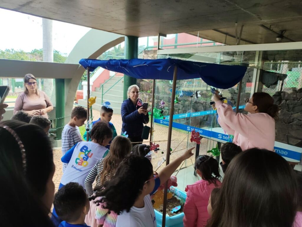 A imagem mostra um ambiente ao ar livre, possivelmente em um parque, zoológico ou centro de educação ambiental, onde um grupo de crianças e adultos está participando de uma atividade educativa. No centro da cena, há uma estrutura com uma cobertura azul, da qual pendem vários peixinhos coloridos feitos de papel ou material leve, presos por fios, formando um tipo de móbile ou jogo interativo. Uma mulher vestindo uma jaqueta rosa está manipulando os peixinhos, enquanto várias crianças, algumas usando camisetas brancas com a palavra "Educação" nas costas, observam atentamente e participam da atividade. Ao lado, outra mulher está fotografando a cena com um celular, registrando o momento. O ambiente é cercado por um muro de pedra e paredes de vidro com a marca "semae", que indica uma possível ligação com uma entidade de saneamento ou meio ambiente da cidade de Piracicaba, conforme também aparece no texto da imagem. A atmosfera geral é de aprendizado e interação, com os participantes envolvidos em uma atividade lúdica que provavelmente tem o objetivo de ensinar sobre meio ambiente, sustentabilidade ou algum tema relacionado à educação ambiental. A iluminação natural sugere que a foto foi tirada durante o dia.