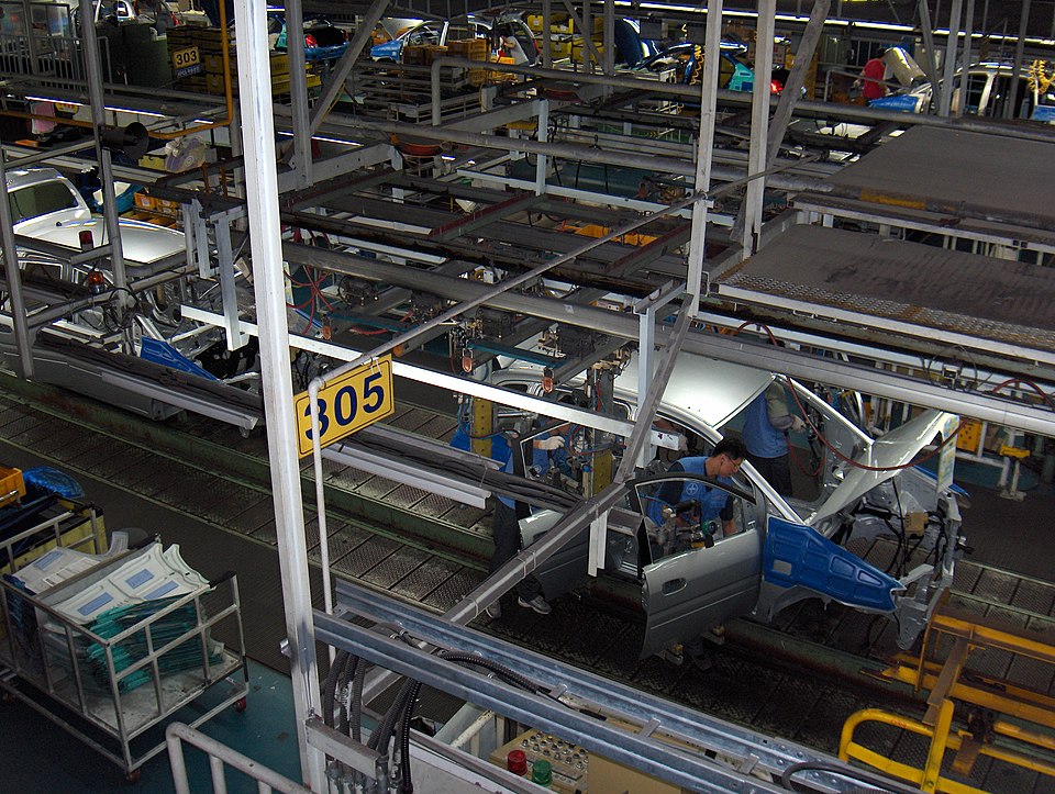 A imagem mostra o interior de uma linha de montagem de uma fábrica automotiva, vista de cima.Detalhes da cena:Local: É uma esteira de produção industrial. Carrocerias de veículos sem pintura final circulam sobre trilhos metálicos.Veículos: Dá pra ver várias carrocerias prateadas e azuis em diferentes estágios de montagem. Uma delas está bem no centro, com as portas abertas.Trabalhadores: Dois funcionários de uniforme azul estão dentro e ao lado da carroceria central, instalando peças. Eles usam EPI e ferramentas pneumáticas.Estrutura: O galpão é cheio de estruturas metálicas, tubulações, cabos e esteiras aéreas. Há placas amarelas de identificação, como a "305" bem visível, e outra "303" ao fundo.Organização: À esquerda tem um carrinho metálico com peças plásticas verdes. Ao fundo, outras estações de trabalho e veículos em processo. O piso é azul industrial.A foto transmite movimento, precisão e trabalho em equipe típico da indústria automobilística. O foco está na montagem manual dentro de um sistema automatizado.