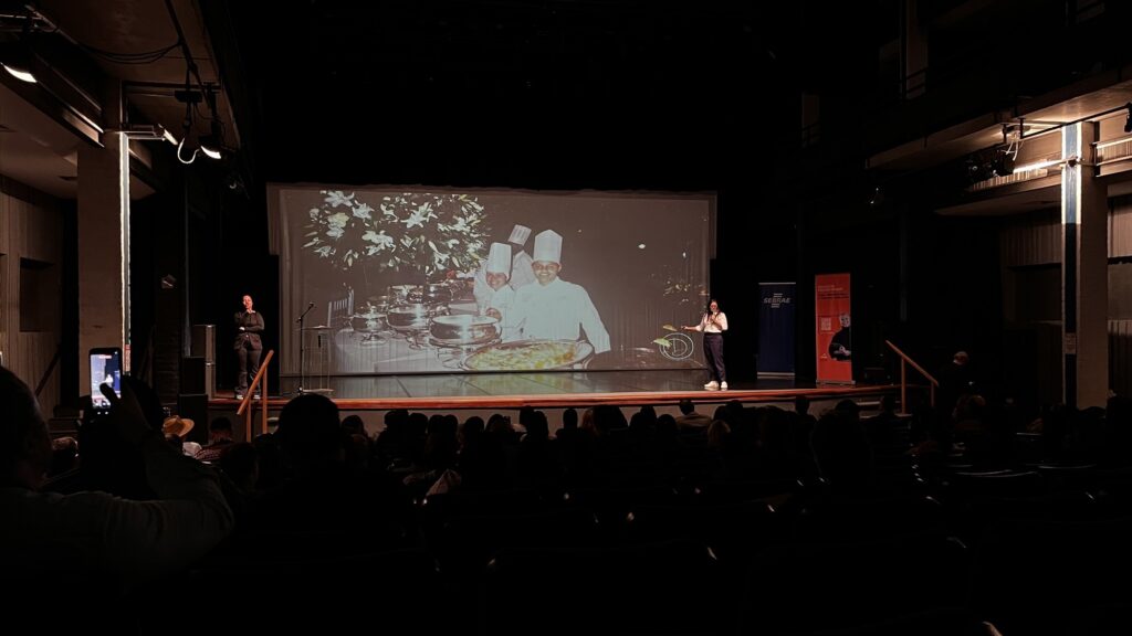 Nesta imagem, vemos um evento ou palestra acontecendo em um auditório com iluminação baixa. No palco, há uma grande tela de projeção exibindo uma foto de dois chefs de cozinha ao lado de uma mesa com pratos preparados e arranjos de flores. Duas pessoas estão no palco: uma mulher à esquerda, vestida de preto, e outra mulher à direita, que parece estar apresentando ou falando ao público. Há banners do SEBRAE nas laterais do palco e uma plateia sentada na parte escura do auditório assistindo à apresentação.