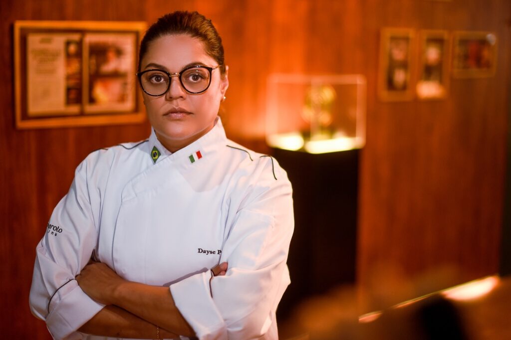 A imagem mostra uma chef de cozinha, Dayse Paparoto, em um retrato médio, com os braços cruzados. Ela usa um uniforme branco de chef, com detalhes que incluem uma pequena bandeira do Brasil e da Itália na gola, além do nome "Dayse P" bordado no peito. Dayse tem cabelos presos para trás e usa óculos de armação redonda. Ao fundo, há uma parede de madeira com algumas molduras penduradas, provavelmente certificados ou prêmios, e uma vitrine iluminada desfocada. A iluminação é quente, destacando a chef e criando um ambiente acolhedor e profissional. A expressão dela transmite confiança e autoridade na área culinária.