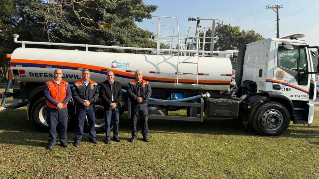 A imagem mostra quatro homens em pé alinhados à frente de um caminhão-pipa branco, que pertence à Defesa Civil do estado de São Paulo, conforme indicado pelas inscrições no veículo ("SP GOV.BR", "DEFESA CIVIL" e "FUNDO SOCIAL SÃO PAULO"). O caminhão tem um grande tanque cilíndrico, uma escada lateral e uma mangueira azul presa na lateral. Os homens estão vestidos com uniformes oficiais da Defesa Civil, exceto um deles que veste um blazer escuro e jeans, sugerindo que ele pode ser um representante ou autoridade. O cenário é ao ar livre, em um campo de grama, com árvores ao fundo sob um céu claro e ensolarado. A iluminação natural destaca o caminhão e as pessoas, criando sombras no chão. A foto foi tirada de um ângulo ligeiramente baixo, o que dá destaque ao tamanho do caminhão-pipa.