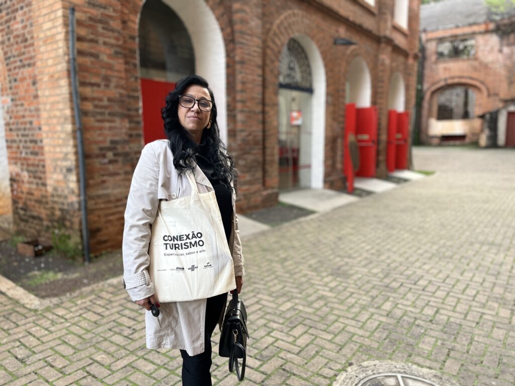 Nesta foto, uma mulher de cabelos pretos, usando óculos de grau e um sobretudo bege, está de pé em um pátio pavimentado com blocos de concreto. Ela segura uma ecobag clara com a inscrição "CONEXÃO TURISMO" e uma bolsa preta. Ao fundo, vê-se uma construção antiga de tijolos aparentes, com arcos brancos e portas vermelhas, o que sugere um ambiente histórico ou cultural.