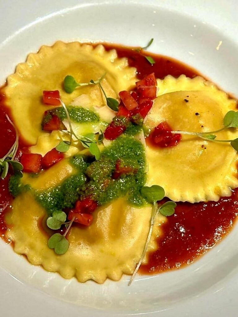 A imagem mostra um prato de massa gourmet, especificamente ravioli, visto de cima. São três raviolis redondos, com bordas levemente onduladas, de cor amarelo-dourada, servidos em um molho vermelho, provavelmente de tomate. Sobre os raviolis, há um molho verde, que parece ser pesto ou algum tipo de azeite temperado, além de pedacinhos pequenos de tomate fresco e algumas folhinhas delicadas de microverdes como decoração. O prato está em uma tigela branca, e a apresentação é limpa e elegante, destacando as cores vibrantes e a textura da massa e do molho. A iluminação é clara, realçando o brilho e o apelo visual da comida.