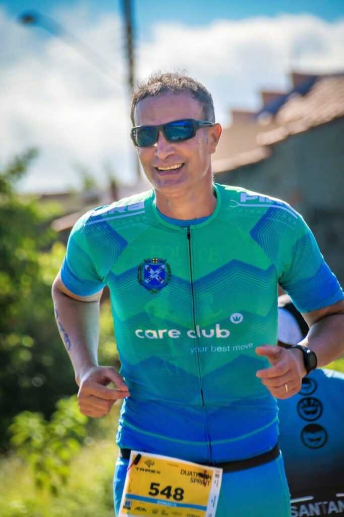 A imagem mostra um homem adulto correndo em um evento esportivo ao ar livre, provavelmente um duatlo. Ele está no centro da foto, da altura do peito para cima, vestindo uma camiseta esportiva de manga curta nas cores verde e azul com detalhes em branco. O homem tem cabelo curto e escuro, está usando óculos escuros esportivos e sorri levemente, demonstrando esforço e satisfação. Na camiseta, há diversos logos, incluindo "care club your best move" e um brasão no lado esquerdo do peito. Ele usa um número de competição preso à cintura com o número 548 e o nome "DONIZETE SANTAN" visíveis, além da inscrição "TAREX DUATHLON SPRINT". O homem também usa um relógio preto no pulso esquerdo. O fundo está desfocado, mostrando vegetação e uma construção, sugerindo que a prova ocorre durante o dia em ambiente externo. A luz natural é intensa, iluminando bem o rosto e a camiseta do corredor. À direita da imagem, é possível ver parte de outro corredor com uma camiseta azul, que tem o logo do banco Santander.